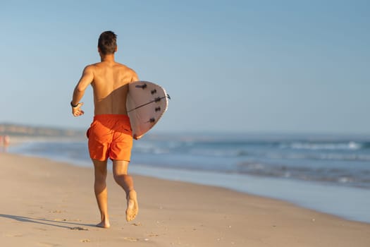 A man with nice athletic body running on the shore holding a surfboard. Mid shot