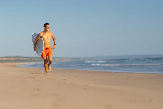 An attractive man with nice athletic body running on the shore holding a surfboard. Mid shot