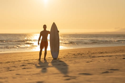 Silhouette of a man standing on the shore holding a surfboard. Mid shot