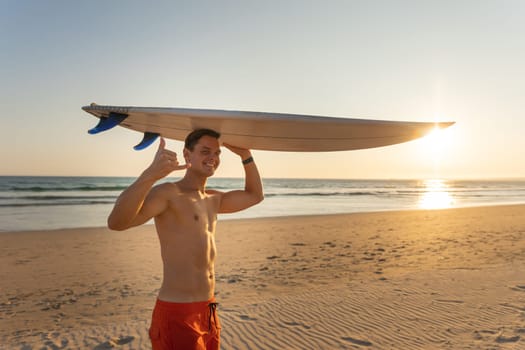 An attractive smiling man with nice athletic body holding a surfboard with one hand and looking in the camera showing shaka. Mid shot