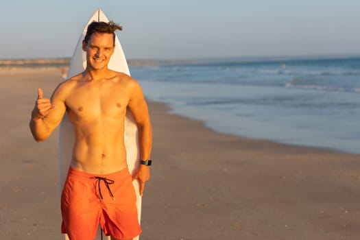 An attractive smiling man with nice athletic body standing on the shore with a surfboard behind his back and showing shaka. Mid shot