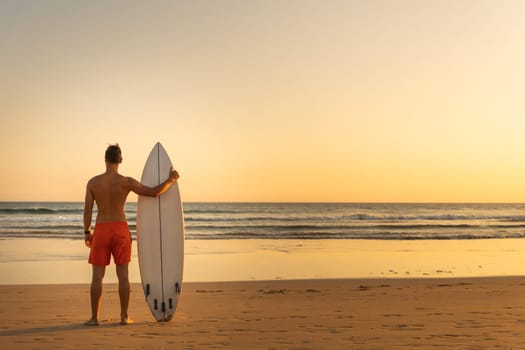 A man standing on the shore holding a surfboard - view from the back. Mid shot