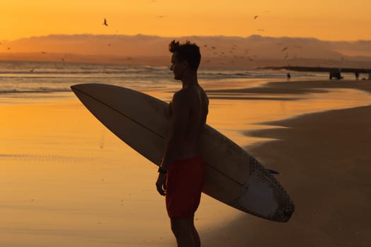 Black silhouette of an athletic man standing on the shore holding a surfboard at sunset. Mid shot