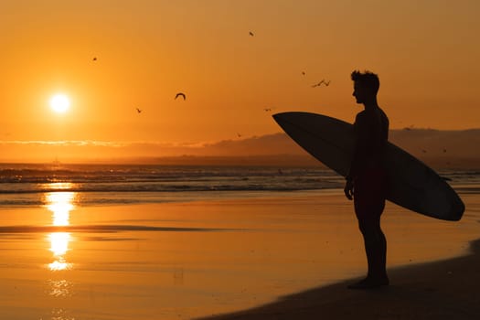 Black silhouette of an athletic man standing on the shore holding a surfboard at orange sunset. Mid shot