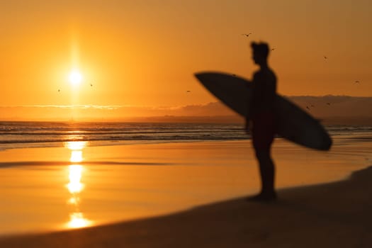 Black silhouette of a man standing on the shore holding a surfboard at orange sunset. Mid shot