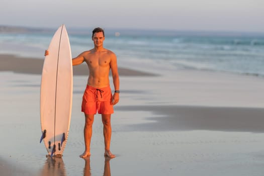An attractive smiling man with nice athletic body standing on the shore holding a surfboard at early sunset. Mid shot