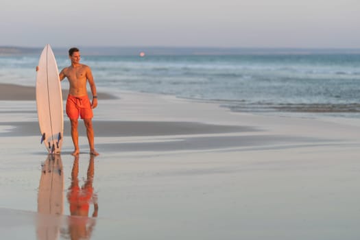 An attractive man with nice athletic body standing on the shore holding a surfboard at early sunset - looking to the side. Mid shot