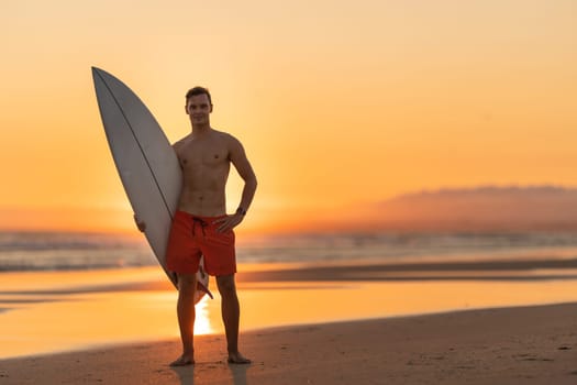 An attractive man standing on the shore holding a surfboard at orange sunset. Mid shot