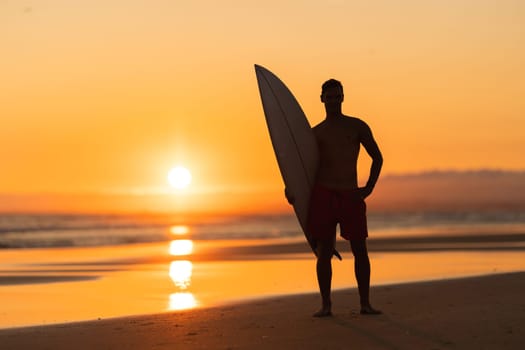 Black silhouette of an attractive man standing on the shore holding a surfboard at orange sunset. Mid shot