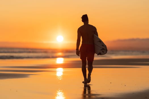 Black silhouette of an attractive man walking on the shore holding a surfboard at orange sunset. Mid shot