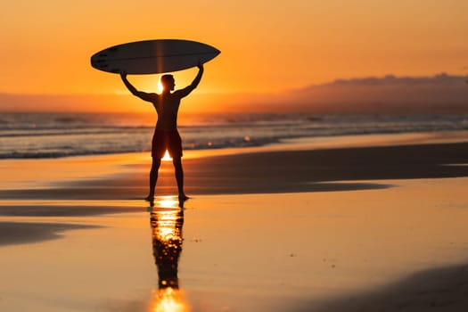 Black silhouette of an attractive man on the shore holding a surfboard above his head at orange sunset. Mid shot