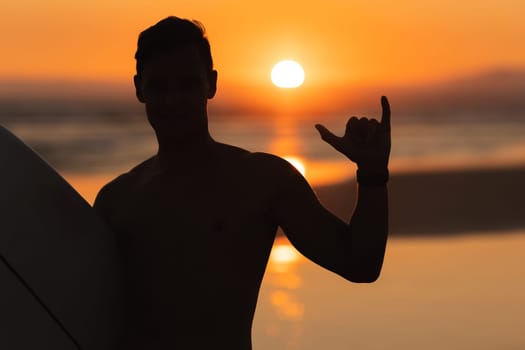Black silhouette of an athletic man surfer showing shaka at sunset. Portrait