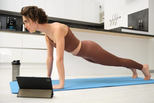 Workout, training and sports concept. Young serious woman follows online training video, stands in plank on rubber yoga mat at home, boosts endurance, excerice on abs.