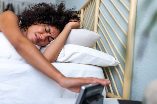 Unhappy, upset and tired African American young woman stopping alarm clock ringing very early in the morning after bad night sleep. Lifestyle concept.