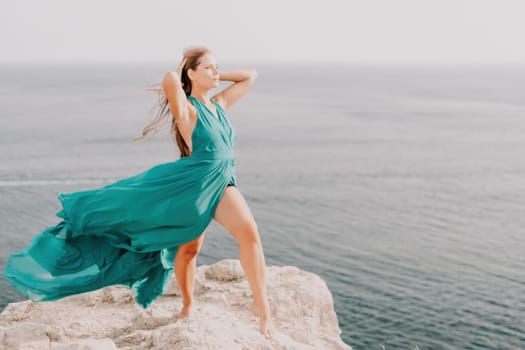 Side view a Young beautiful sensual woman in a mint long dress posing on a volcanic rock high above the sea during sunset. Girl on the nature on overcast sky background. Fashion photo