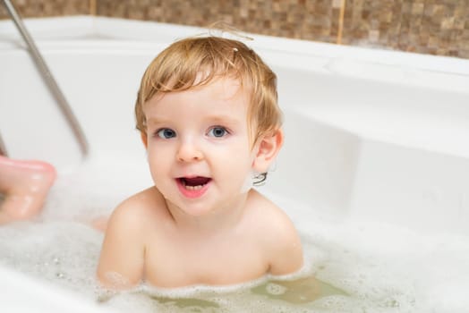 Funny little boy getting a bath in the bathroom. A child bathes in water with soap suds in white bath.