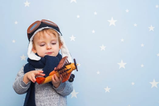 Little boy in helmet and glasses with wooden plane against the background of a blue wall with stars. Happy child playing with toy airplane.