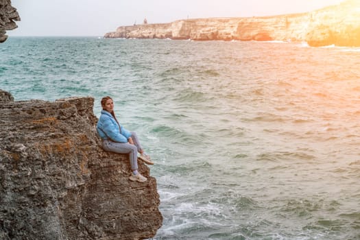 woman sea travel. A woman in a blue jacket sits on a rock above a cliff above the sea, looking at the stormy ocean. Girl traveler rests, thinks, dreams, enjoys nature.