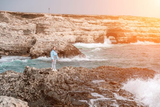 woman sea travel. A woman in a blue jacket stands on a rock above a cliff above the sea, looking at the stormy ocean. Girl traveler rests, thinks, dreams, enjoys nature.