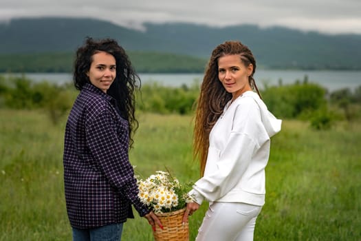 Two women enjoy nature in a field of daisies. Girlfriends hugging hold a bouquet of daisies and look at the camera