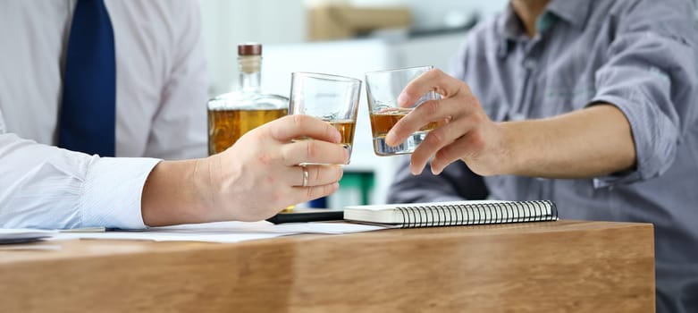 Close-up of hands businessmen holding glasses with alcohol drinks. Partners celebrating signing profitable contract in office. Business negotiations concept