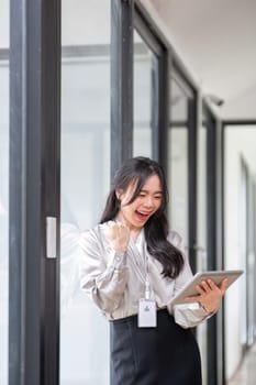 A beautiful and cheerful Asian businesswoman or female boss is showing her clenched fist and looking at her tablet with an excited face, receiving good news and achieving goals.