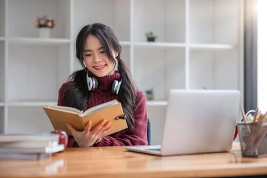 Asian female student studying online using headphones and laptop taking notes in notebook sitting at home table relaxing and changing places to school relax..