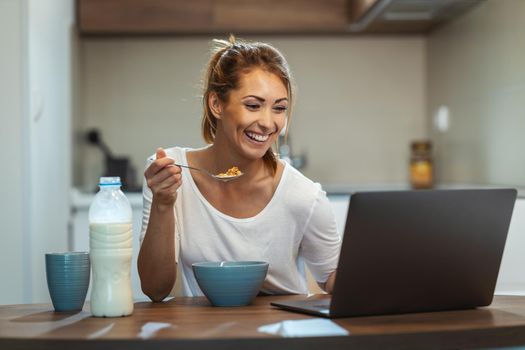 Beautiful young woman is eating her healthy breakfast in her kitchen and using laptop to make a video chat with someone.