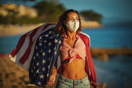 Attractive young woman with US national flag and protective N95 mask on the beach during preventing the spread of the epidemic and treating coronavirus and pandemic COVID-19.