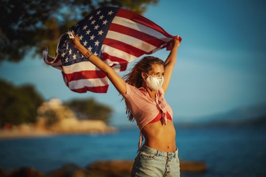 Attractive young woman with US national flag and protective N95 mask on the beach during preventing the spread of the epidemic and treating coronavirus and pandemic COVID-19.