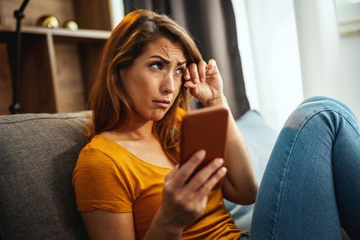 A unhappy young woman is sitting on the sofa and using smartphone to surfing social media at home.