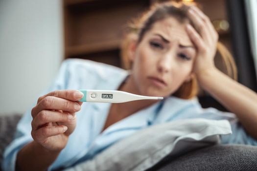 Young woman is lying sick at home couch and looking at thermometer.