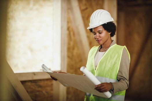 Shot of an African female architect checking blueprints at the construction site of a new wooden house. She is wearing protective workwear and white helmet.