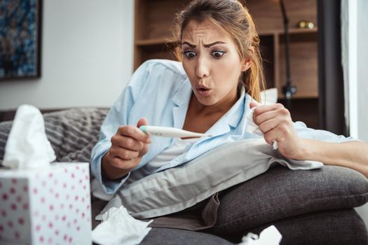 Young woman is lying sick at home couch and looking at thermometer.