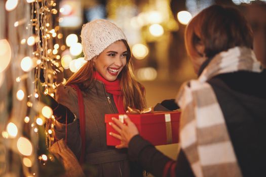 Cheerful two young women are having fun in the city street at the Christmas night. They are laughing and giving holidays presents to each other.