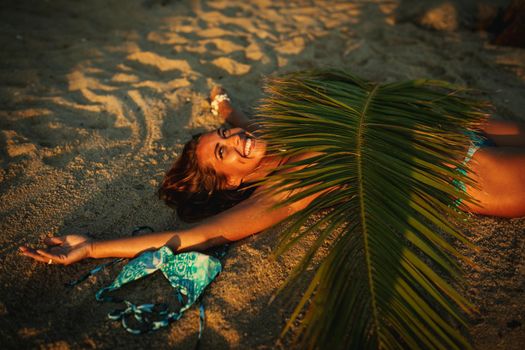 Shot of an attractive young woman is lying on the tropical sandy beach covered with palm tree leaf.