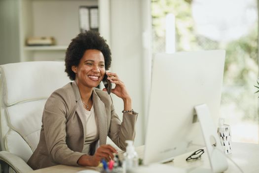 Shot of an African businesswoman talking on smartphone while working at desk in her home office during corona virus pandemic.