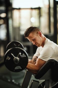 Shot of a muscular guy in sportswear working out at the hard training in the gym. He is pumping up biceps muscule with heavy weight.