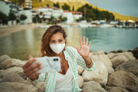 Shot of an attractive happy young woman wearing a protective N95 mask and making video call with her smartphone while spending time on the seaside during the COVID-19.