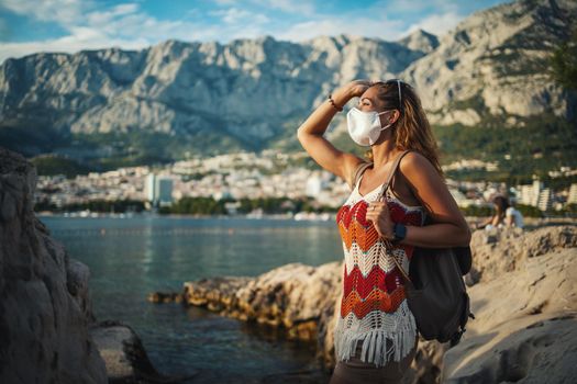 Smiling young female turist with protective mask spending time on seaside during exploring a Mediterranean at corona pandemic. 