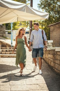 A beautiful young couple is having fun while walking around a Mediterranean town. They are enjoying in summer sunny day, holding each other's hand and smiling.