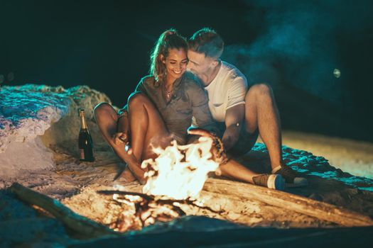The happy smiling couple is sitting on the sandy seashore by the fire in the evening. They are frying sausages on stick.