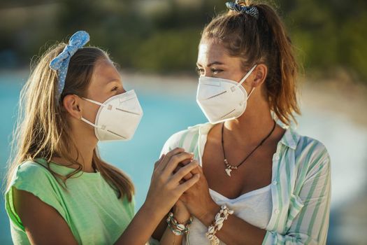 Shot of a smiling sisters with protective N95 mask spending time on the beach at corona pandemic. They are having self-isolation in nature.