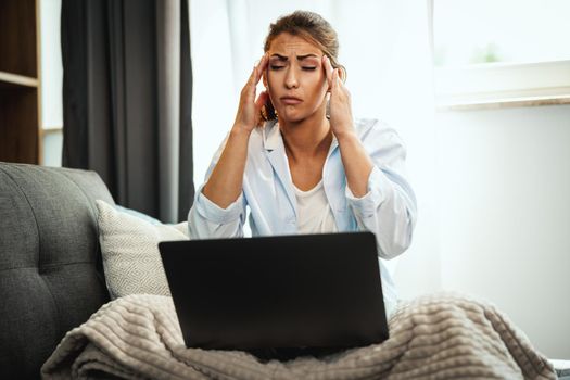 A worried young woman is sitting cross legged on the sofa and using her laptop at home.