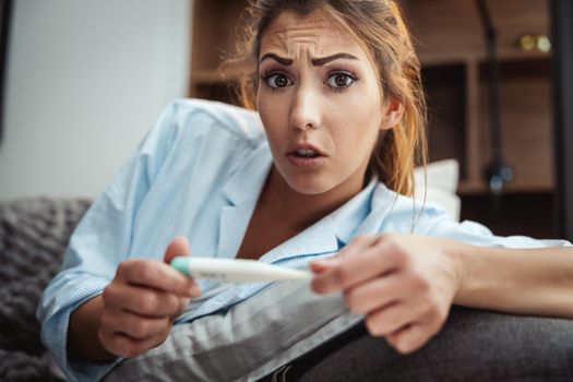 Young woman is lying sick at home couch and looking at thermometer.