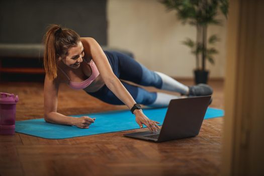 Young smiling woman is doing fitness exercises in the living room on floor mat and looking at the laptop, in morning sunshine.