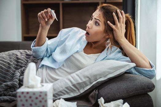 Young woman is lying sick at home couch and looking at thermometer.