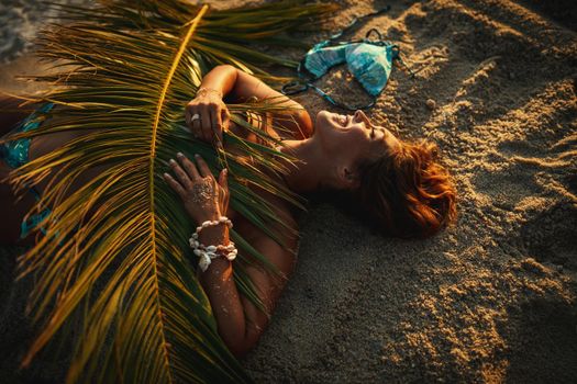 Shot of an attractive young woman is lying on the tropical sandy beach covered with palm tree leaf.