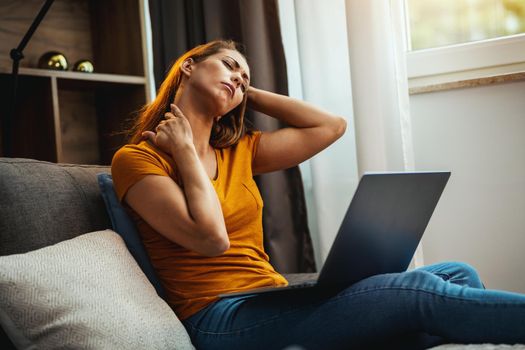 Tired young woman experiencing stress and holding her neck in pain while sitting on the sofa and using her laptop at home.