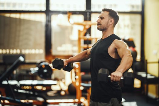 Shot of a muscular guy in sportswear working out with dumbbells at the cross training gym. He is pumping up shoulders muscule with heavy weight.